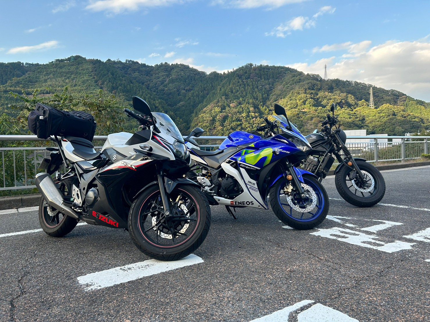3台を側面から。手前のスズキと奥のネイキッドにはキャンプ用と思われる大型バッグが積まれていた。青空と駐車スペースの白線